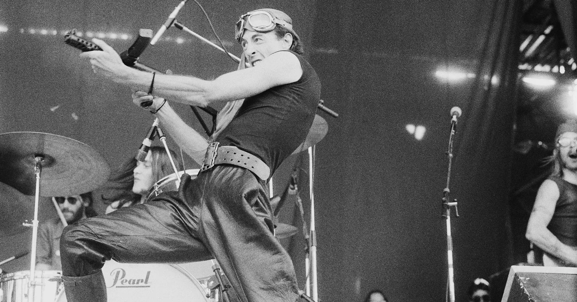 Black and white photo capturing a dynamic live performance moment of Robert Calvert from Hawkwind, known for his theatrical stage presence. He is energetically playing a guitar and wearing a pilot's cap and goggles, embodying his unique rock persona. The drummer in the background adds to the energetic ambiance, highlighted by the dramatic lighting of the stage