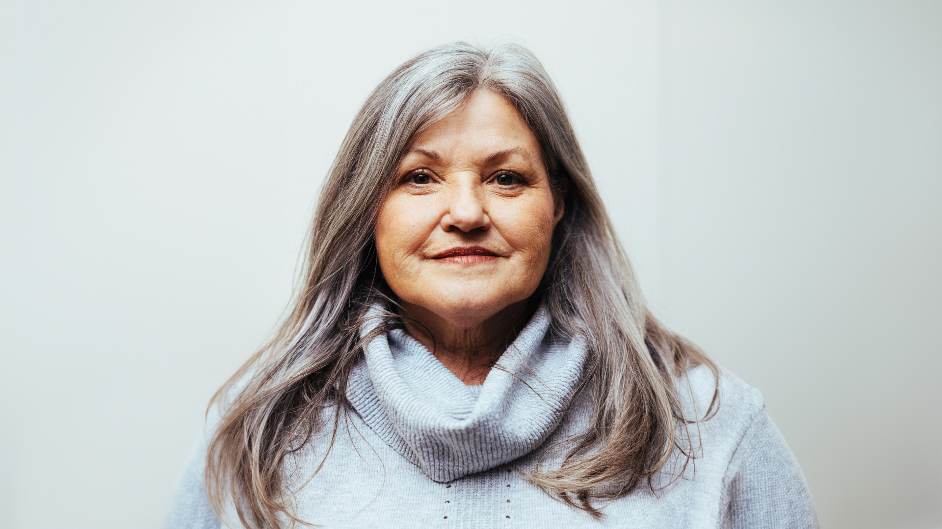 Close-up portrait of Bev Standing, a distinguished voice actress, featuring her serene expression. She has long, silver hair and wears a light blue turtleneck sweater, symbolizing her warmth and approachable nature. The neutral background highlights her contemplative gaze, reflecting her extensive experience and professionalism in the voice acting industry