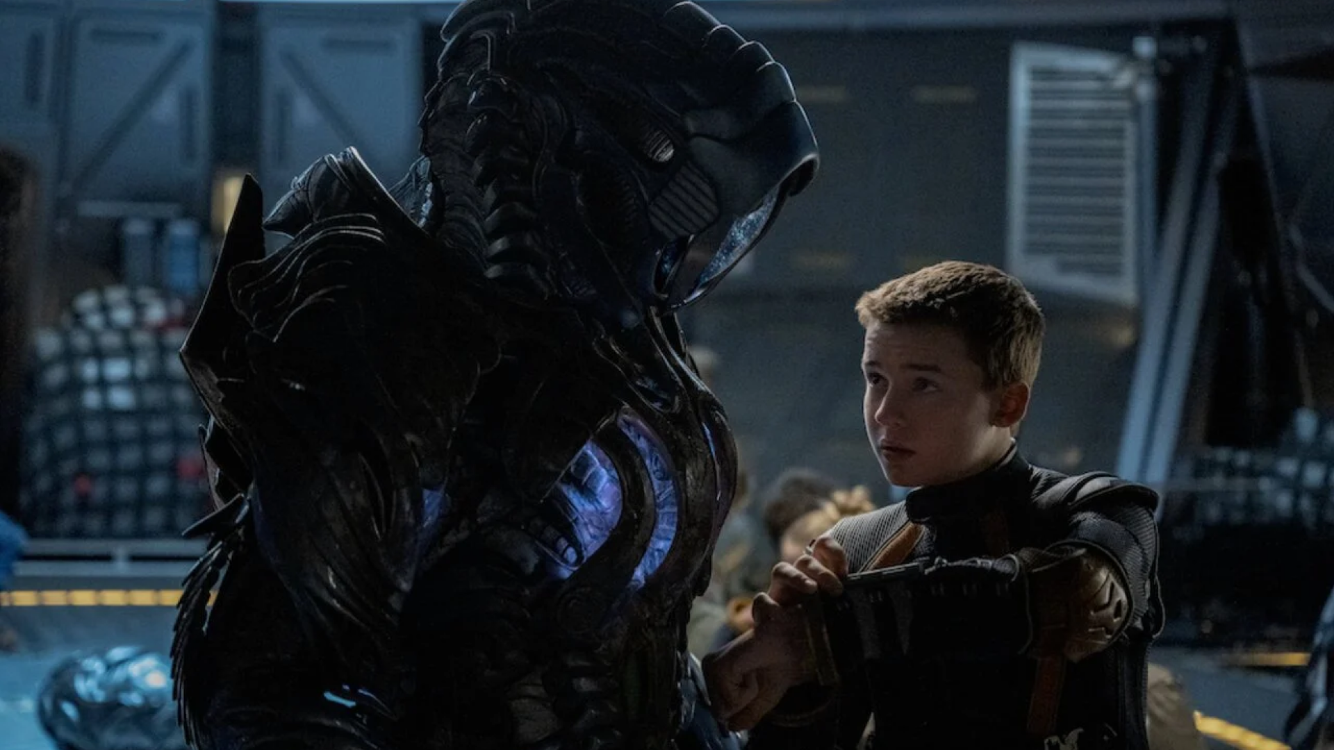 Image featuring a dramatic scene from a science fiction movie featuring a young boy in a space suit facing a menacing robot. The robot, depicted with intricate black armor and a helmet-like head, towers over the boy in a dimly lit spacecraft setting. The tension between the characters is palpable, illustrating a pivotal moment of interaction.