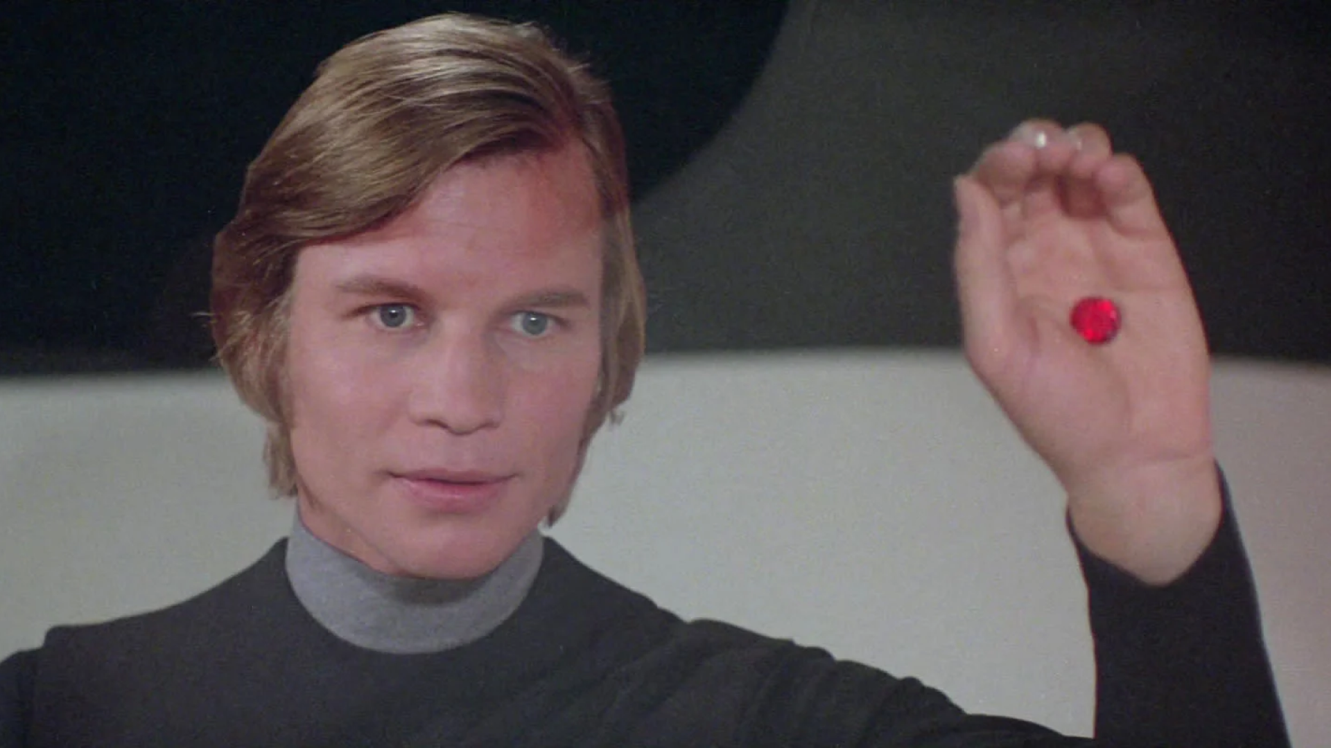 Michael York holding a red object in a promotional image for a healthcare initiative. The actor is dressed in a dark futuristic outfit, his expression calm and focused as he offers the pill, symbolizing choice or a significant decision. The setting is minimalistic with a dark background, emphasising the pivotal moment