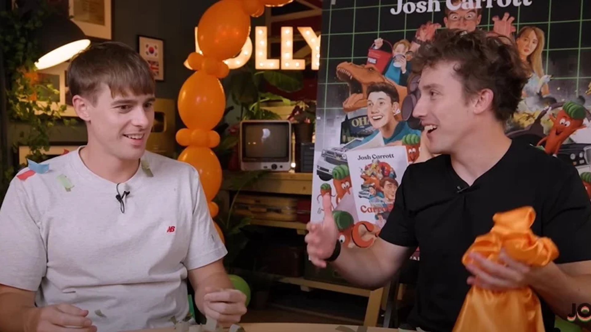 Two young male YouTubers from Jolly channel excitedly engaging in conversation at a vibrant and creatively decorated set with orange balloons and character posters. One wears a grey t-shirt and the other a black t-shirt, both animatedly discussing over a table in a casual, friendly atmosphere