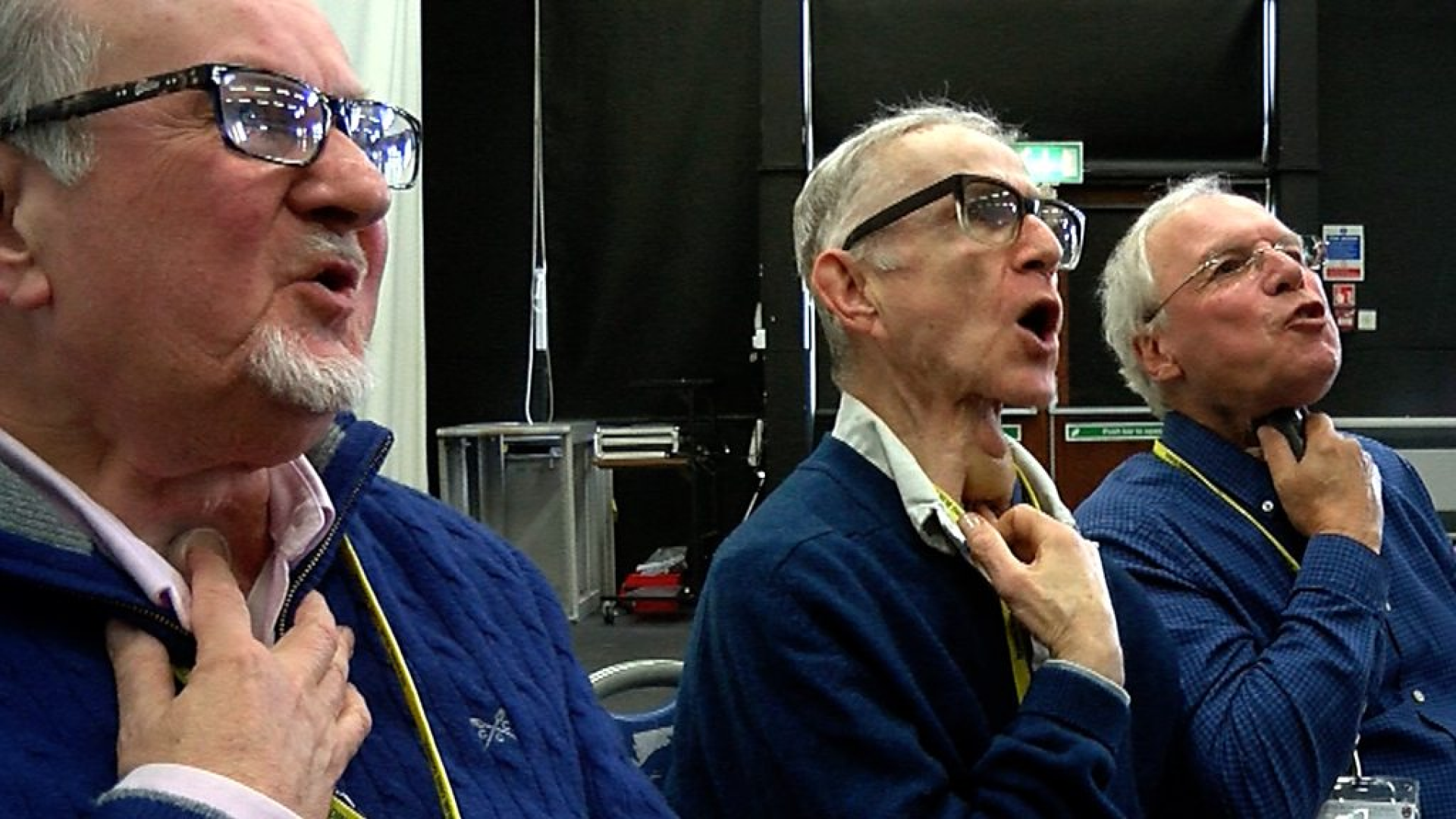 Three elderly men with voice prostheses engaging in vocal exercises at a rehabilitation session. Each man is seen adjusting his neck device, actively participating in voice recovery training in a well-equipped medical setting