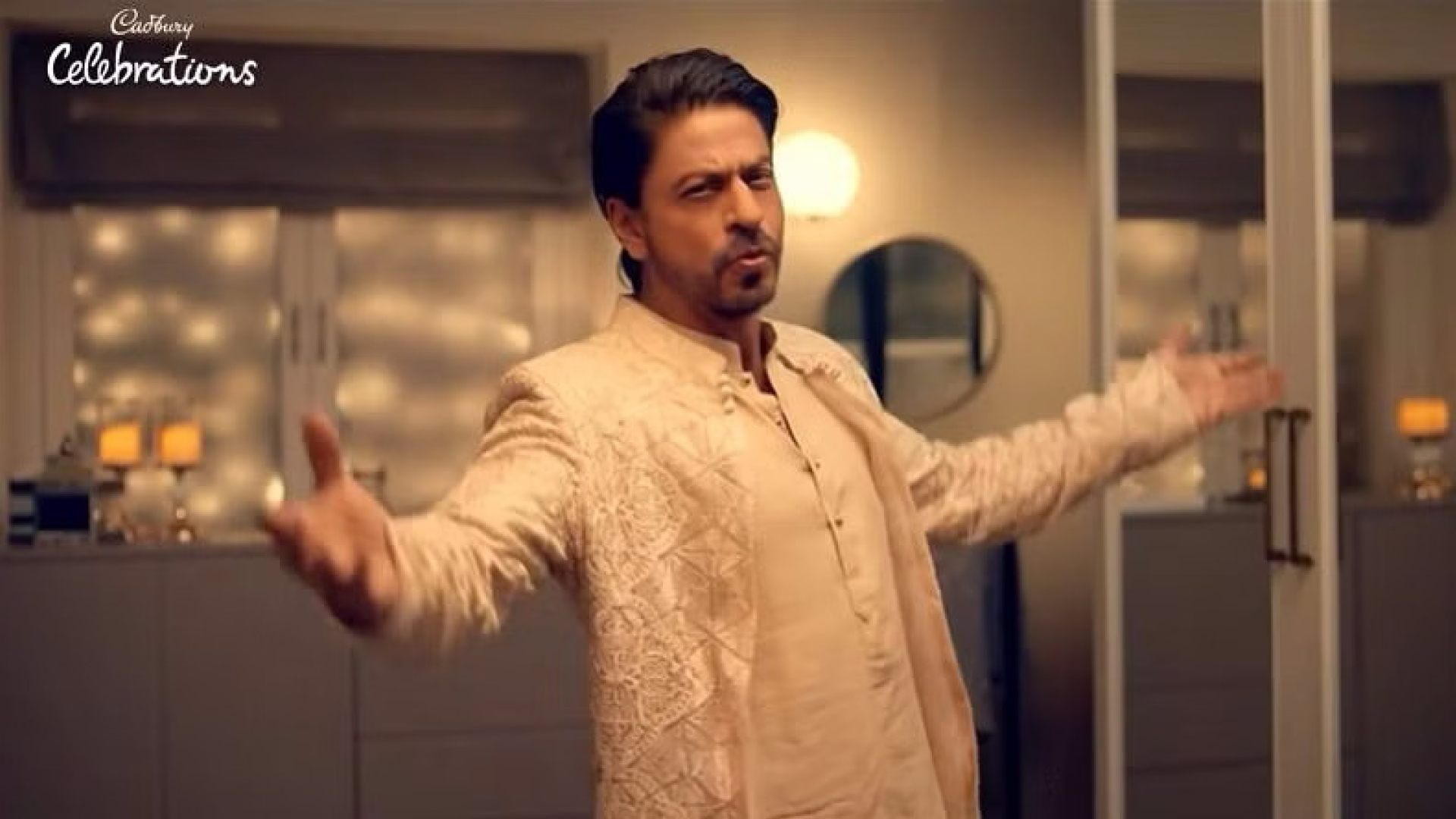 Promotional image of Shahrukh Khan in a Cadbury Celebrations advertisement, joyfully raising his arms in a celebratory gesture. He is dressed in a traditional cream-colored kurta with intricate embroidery, exuding charisma and elegance in a warmly lit indoor setting. The Cadbury Celebrations logo is subtly placed, enhancing the festive and luxurious ambiance of the advertisement