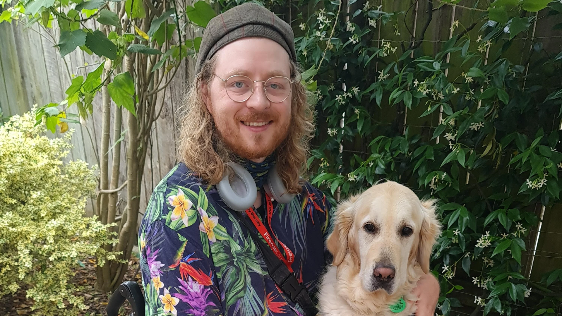 A cheerful man with long curly hair, wearing a tropical shirt and glasses, smiling as he holds a golden retriever in a lush garden setting. The man, accessorized with headphones around his neck and a lanyard, enjoys a sunny day outdoors with his friendly dog