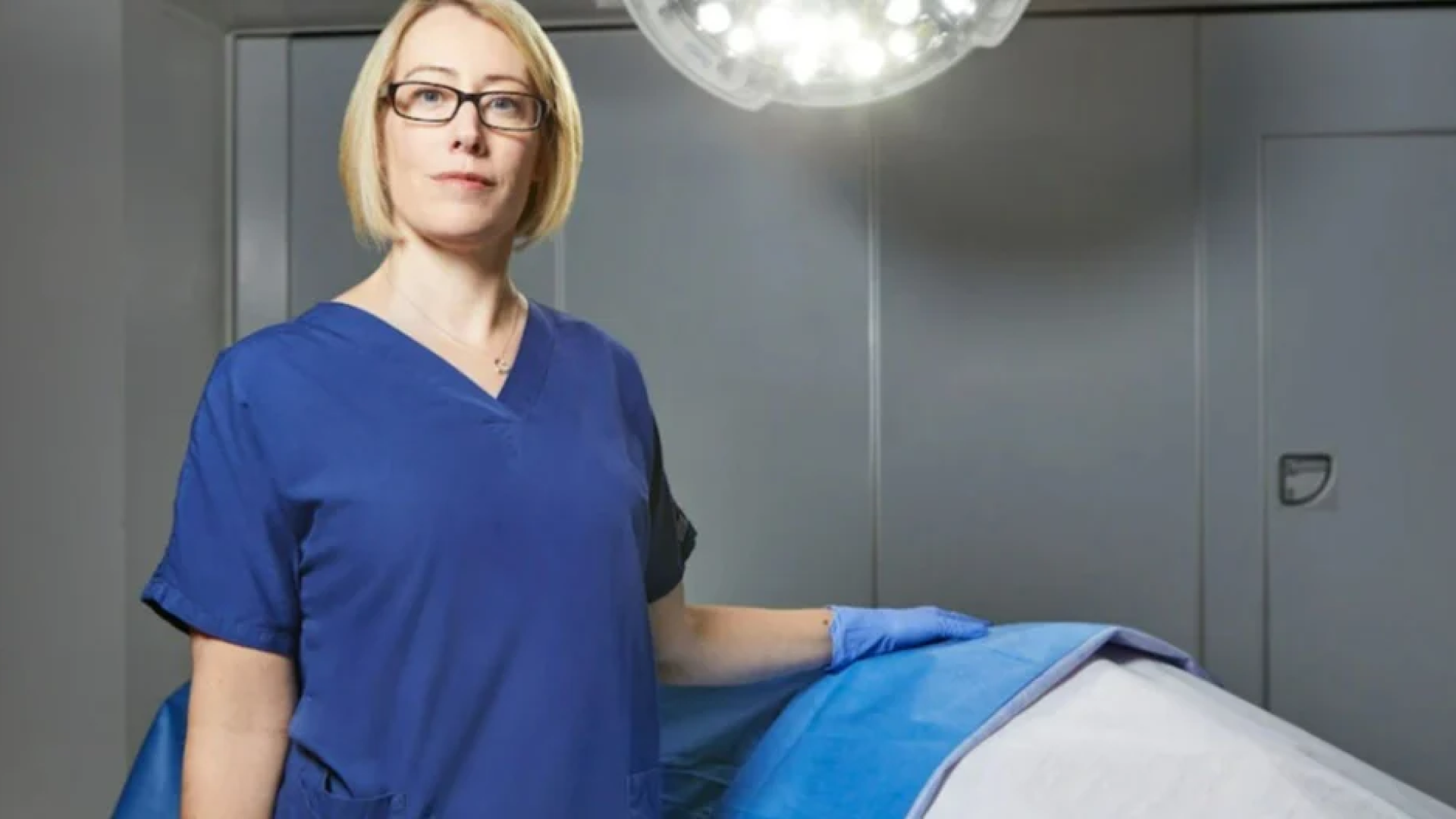 Confident female medical professional in a blue scrub suit standing beside a surgery table in a well-lit operating room. She appears prepared and attentive, ready for a medical procedure, highlighting professionalism and care in healthcare settings