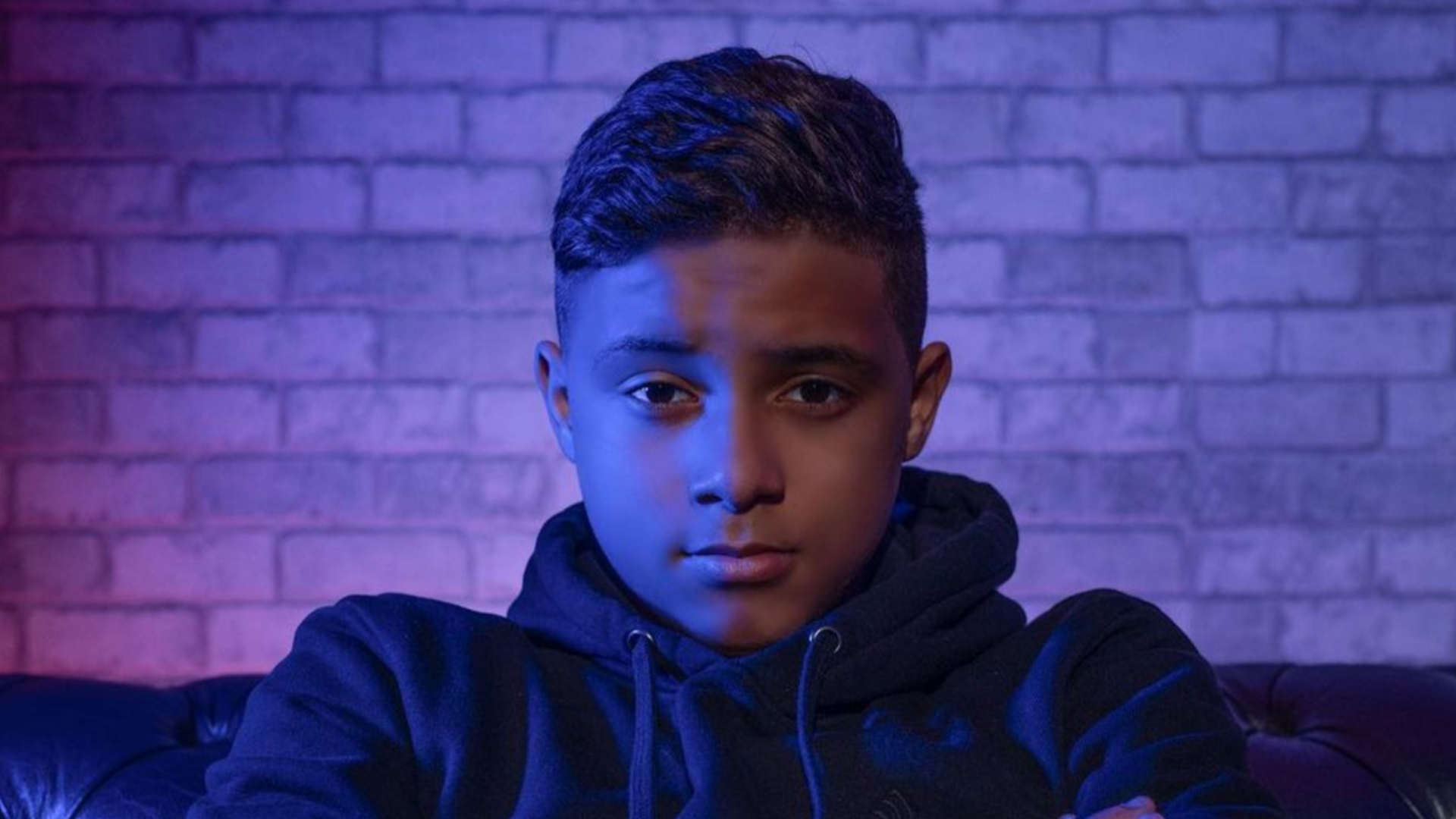 Portrait of a young boy in a dark hoodie sitting against a brick wall under blue and purple lighting, showcasing a serious expression, featured in the promotional material for '10 Years 10 Ans,' a French-Mauritian comedy highlighting Respeecher's voice synthesis technology