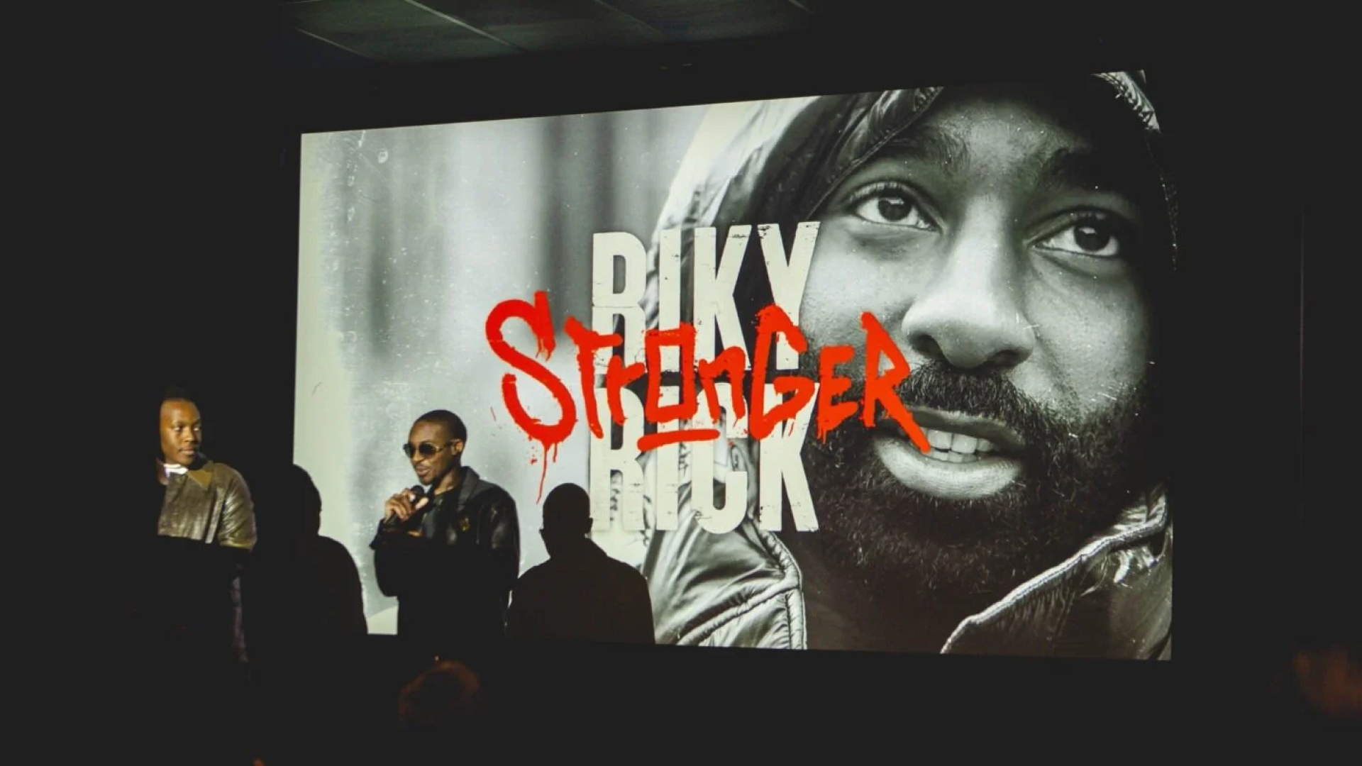 Image featuring a large projection of Riky Rick with the title 'RIKY RICK: STRONGER' in bold, red, graffiti-style text, showcasing a close-up of his face looking introspective