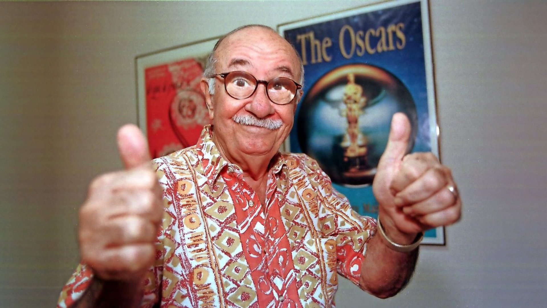 Cheerful elderly actor Tommy Muñiz in a vibrant patterned shirt giving two thumbs up in front of an 'Oscars' poster, symbolising enthusiasm and approval in entertainment.