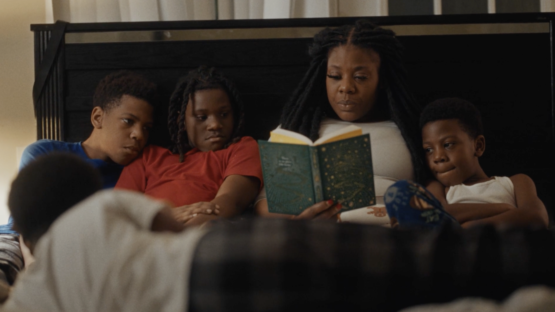 A cozy family reading scene with a mother and three children in bed. The mother, with long dreadlocks, holds an illustrated book open, captivating her young audience. To her left, two boys in blue and red shirts look on attentively, while a third child in a white tank top smiles, enjoying the story.